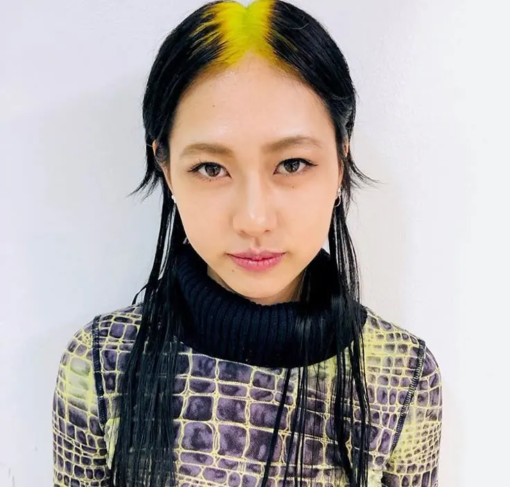 Straight black hair with neon yellow ghost roots and a sharp center part.